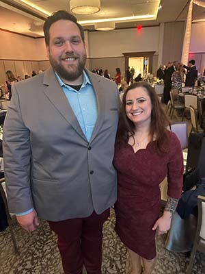 Petite woman wearing an eggplant colored dress smiles while standing with a tall man.