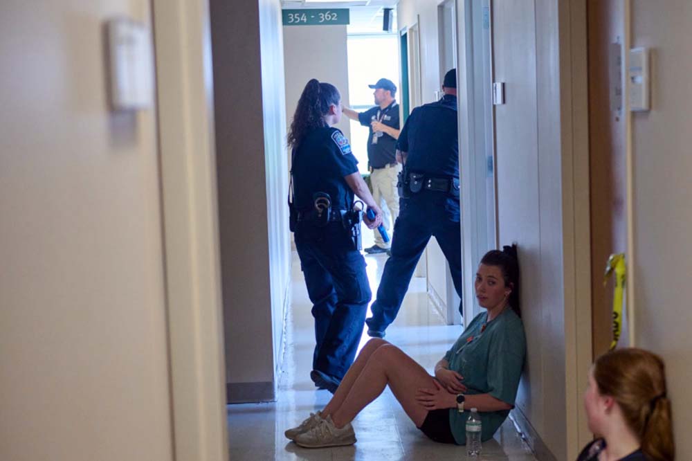 Two young woman sit in a hallway. Behind them are three uniformed officers looking around.