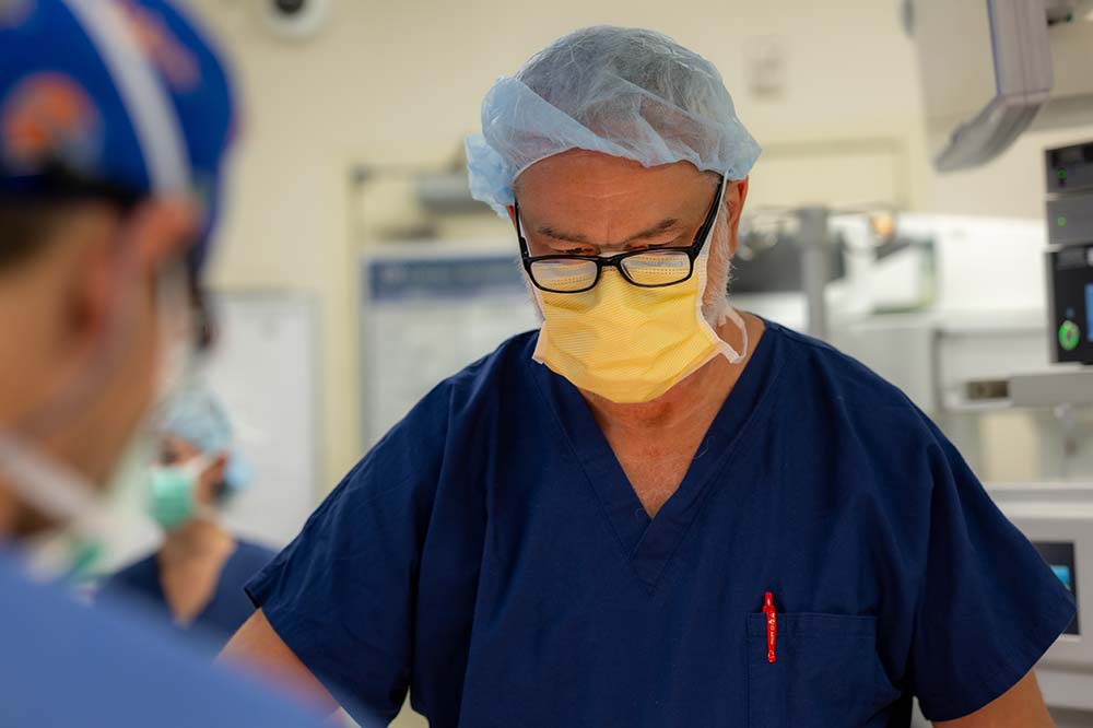 Man wearing blue scrubs, a yellow surgical mask, glasses and a hair cover looks down at an operating table.
