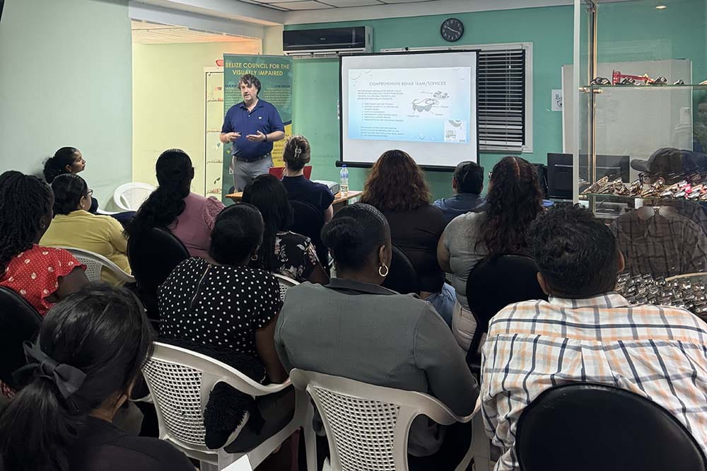 A man wearing a blue shirt standing by a screen speaks as people facing him listen.