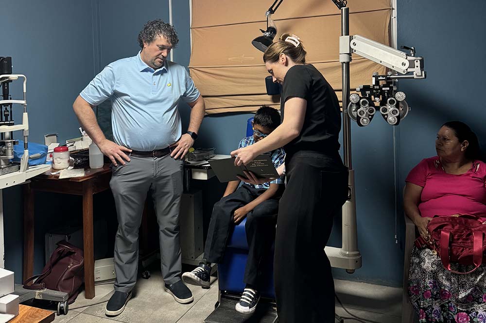 Man wearing a light blue shirt and gray pants talks with a seated patient. A woman wearing dark colored scrubs is on the right, as is another woman who is seated.