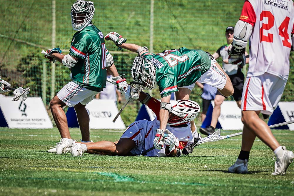 Three lacrosse players. Two are wearing green, one is wearing blue. the one in blue is on the ground as one of the green players appears poised to fall over him.