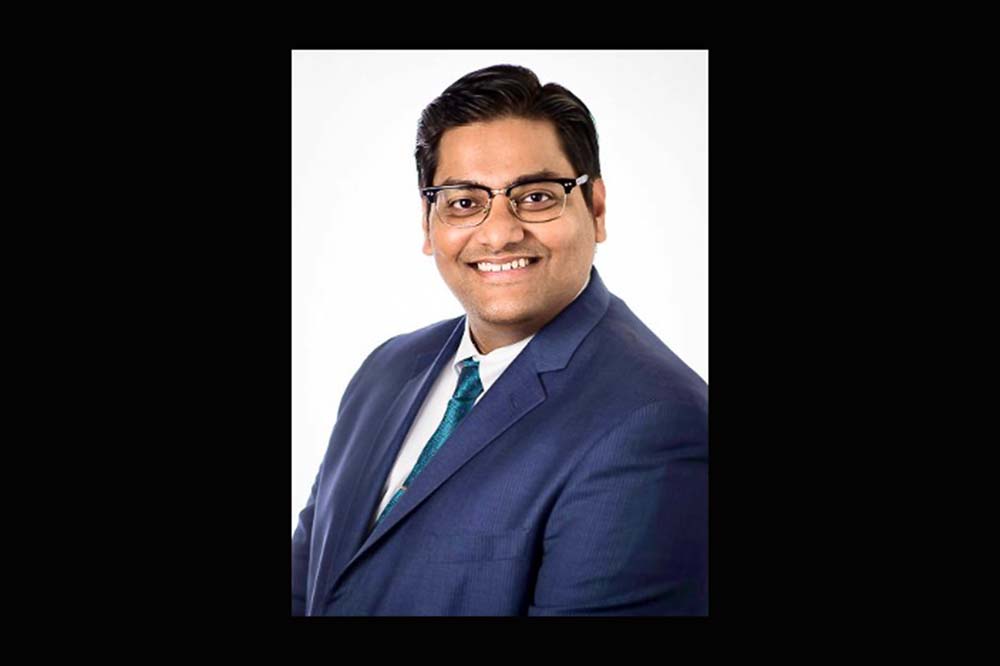 Smiling man wearing glasses, a blue suit coat and a tie.