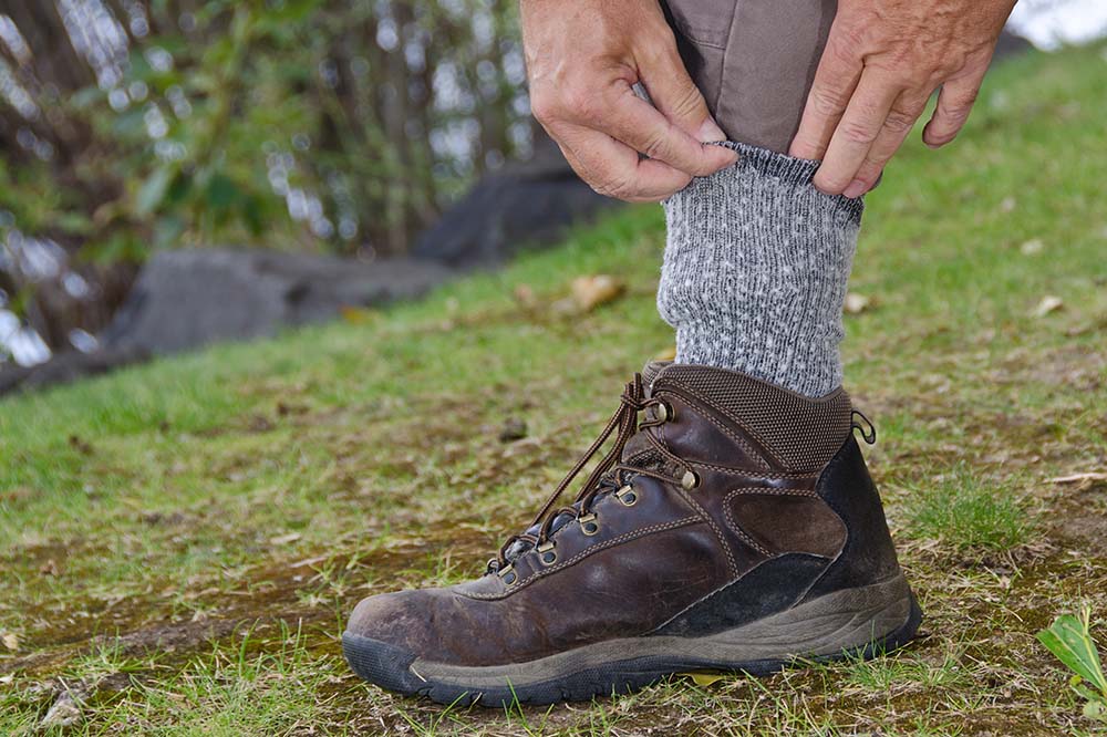 Two hands tuck a brown pants leg into a gray sock. The person is wearing a brown hiking boot. 