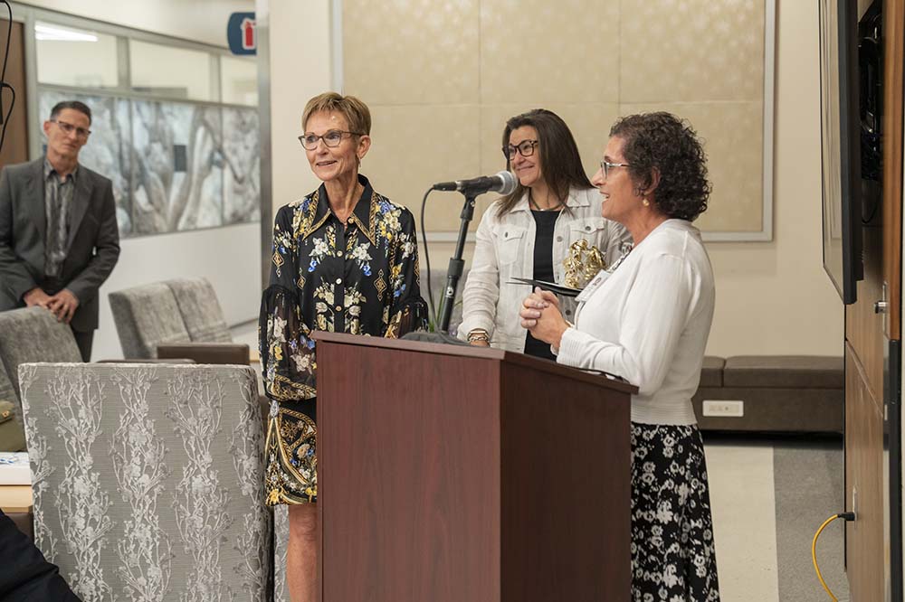 Three women stand behind a podium. The one on the far left has short, light colored hair and is wearing glasses and a dark colored dress. The one in the middle also wears glasses. She has dark hair and is wearing black and white. The third woman has dark curly hair and is wearing a white sweater.