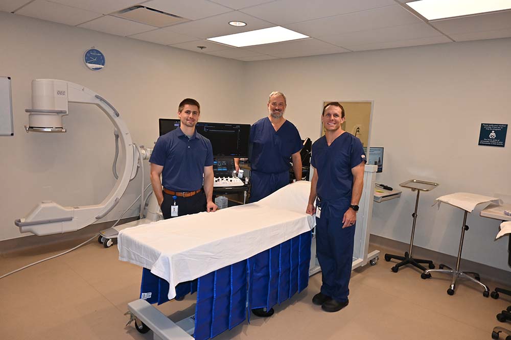Three men wearing blue surgical scrubs stand beside a table draped in white.