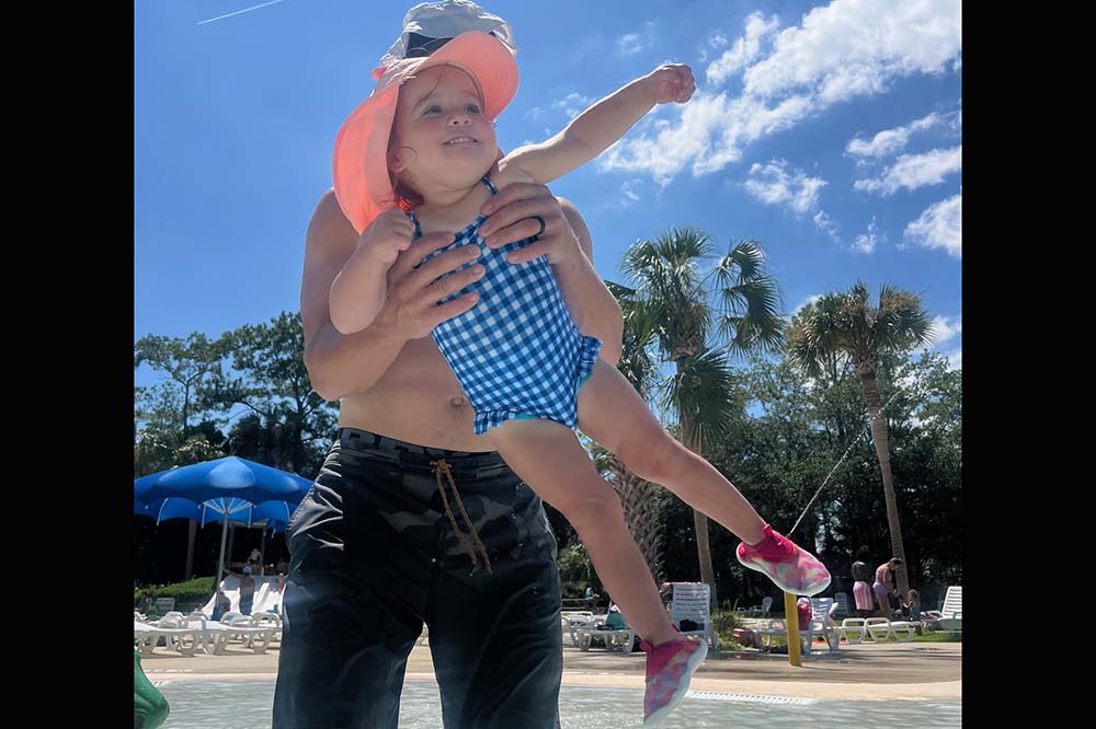 A little girl in a blue checked swimsuit and pink hat smiles and reaches up as her father holds her.
