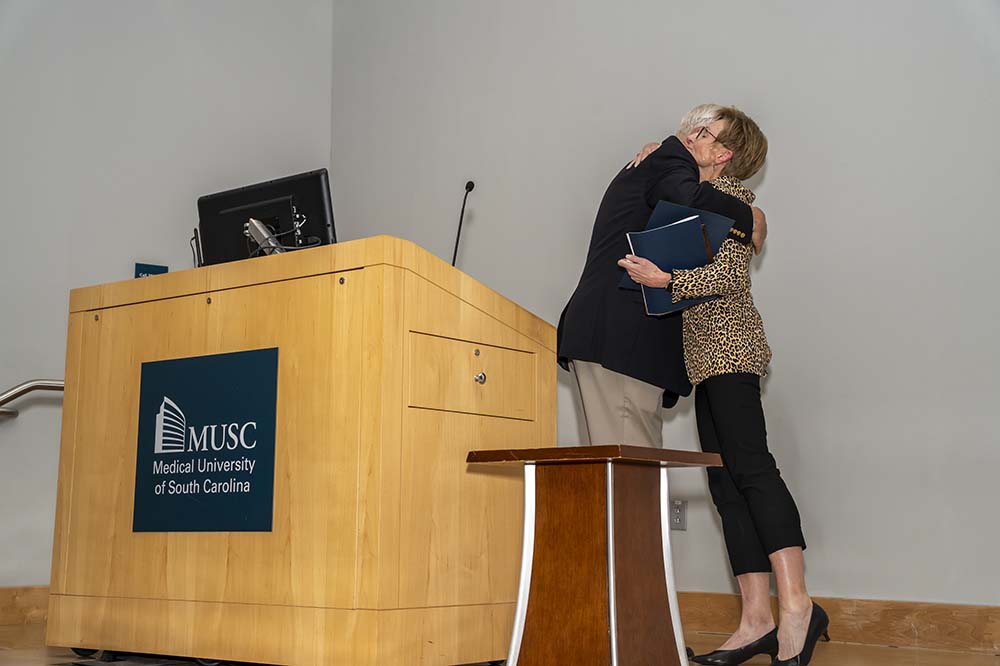 A tall man hugs a woman. They are behind a podium.