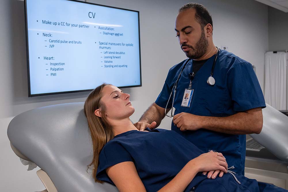 A woman lies on a medical chair while a man examines her. Both are wearing blue scrubs.