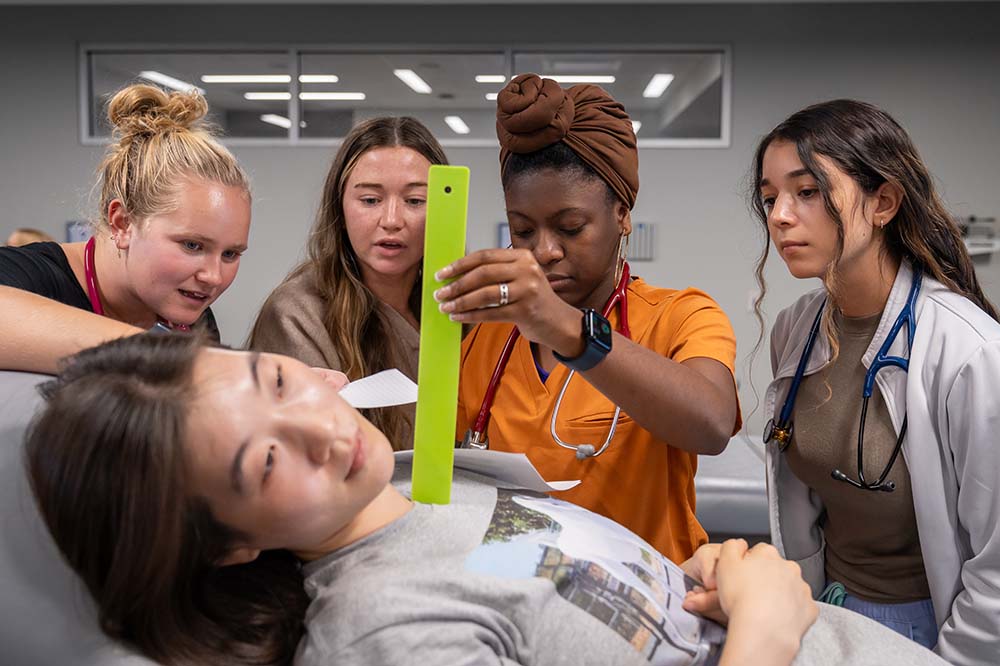 Four young women are standing in front of a fifth woman who is lying down. One is holding a bright yellow stick to the chest of the woman who is lying down.