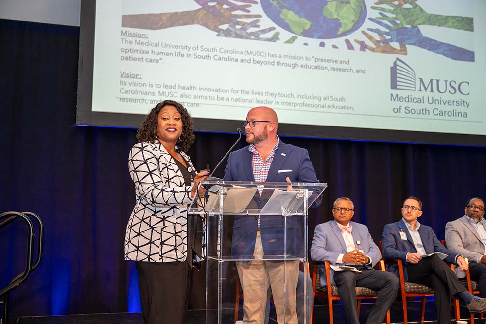 A woman wearing a white jacket with a black design on it stands beside a man wearing business clothing. They are on a stage with a slide behind them.
