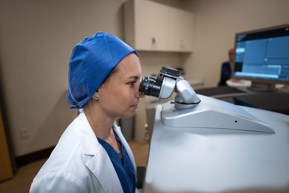 Woman wearing a blue head covering and white doctor's coat lean forward as she looks into a machine.
