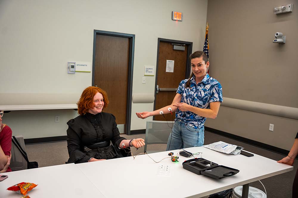 A student wearing black sits at a table. There are wires leading to electrodes on the student's arm. A woman stands next to her, gesturing with her right arm.