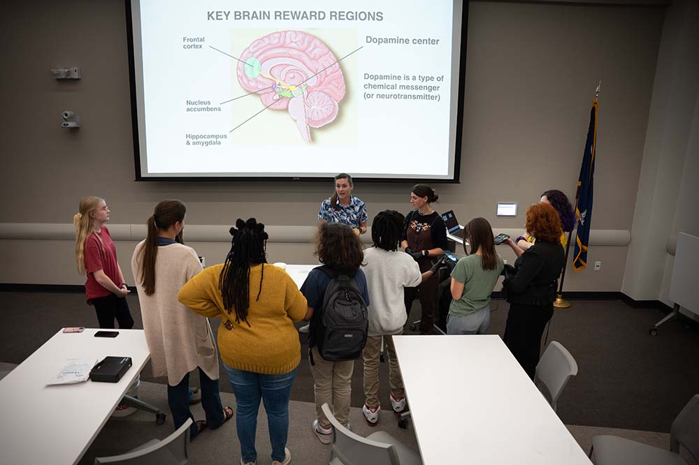 Several students face a woman who is standing in front of a large screen. It shows an image of a brain with the words Key Brin reward regions. Dopamine center. Dopamine is a type of chemical messenger Hippocampus and amygdala. Nucleus accumbent. Frontal cortex.