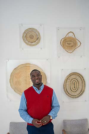 A smiling man wearing a red sweater vest and blue shirt stands in front of sweetgrass baskets hanging on a wall.