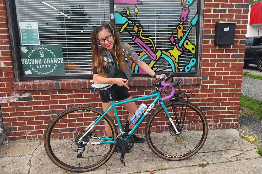 a woman poses with a bike in front of a bike shop