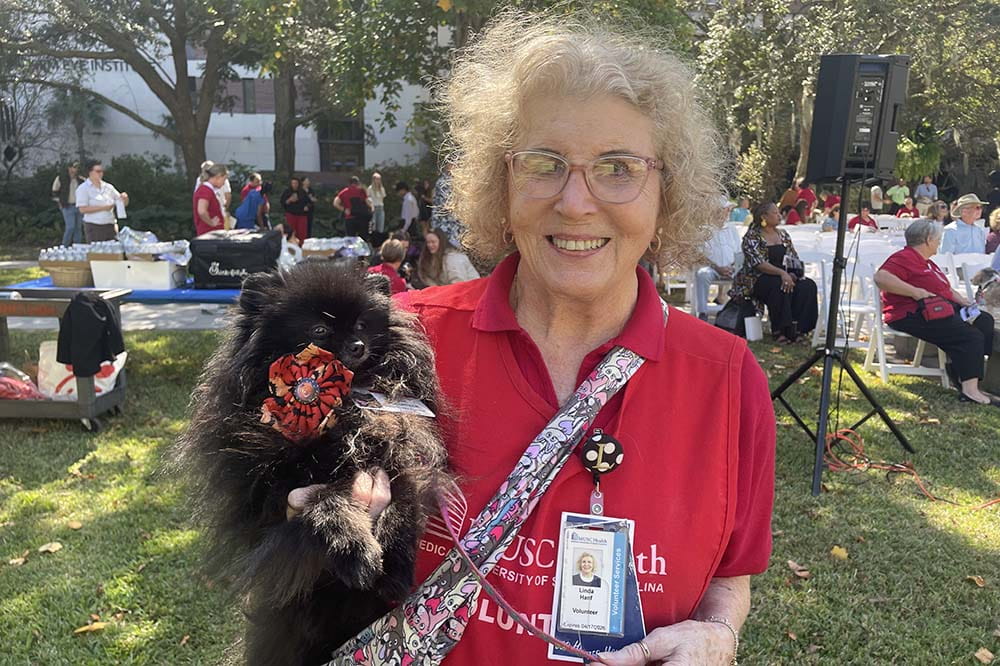 A woman wearing a red shirt holds a small black dog. They are outside at an event.