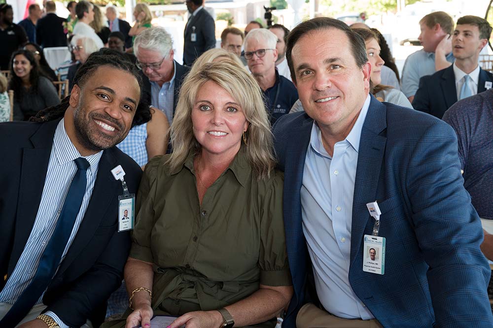 Two men and a woman smile while they're seated. All are wearing business clothing. There are people seated behind them.